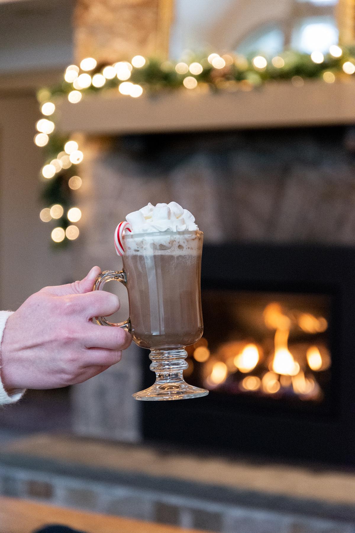 Hand holding a whipped cream-topped hot chocolate by a glowing fireplace.