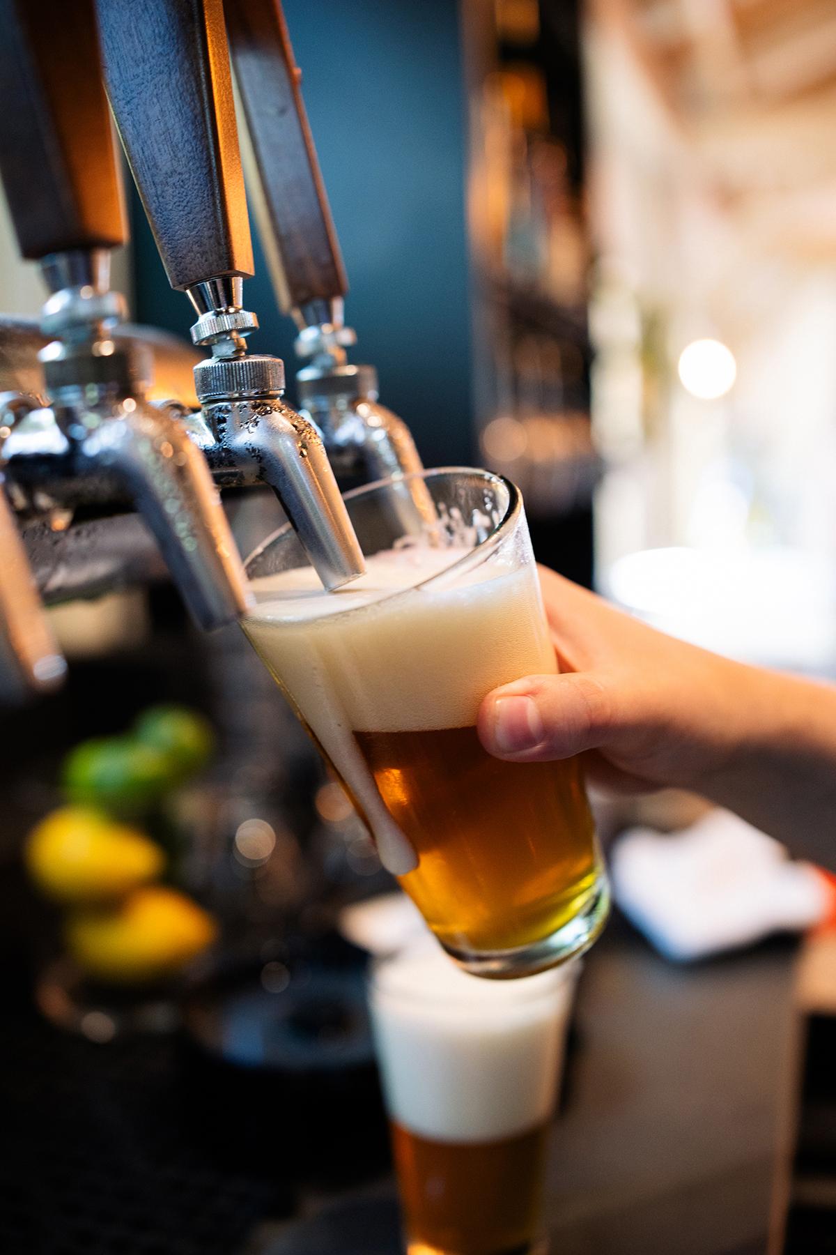 Hand holding a tilted beer glass under taps, filling with amber beer and foam.