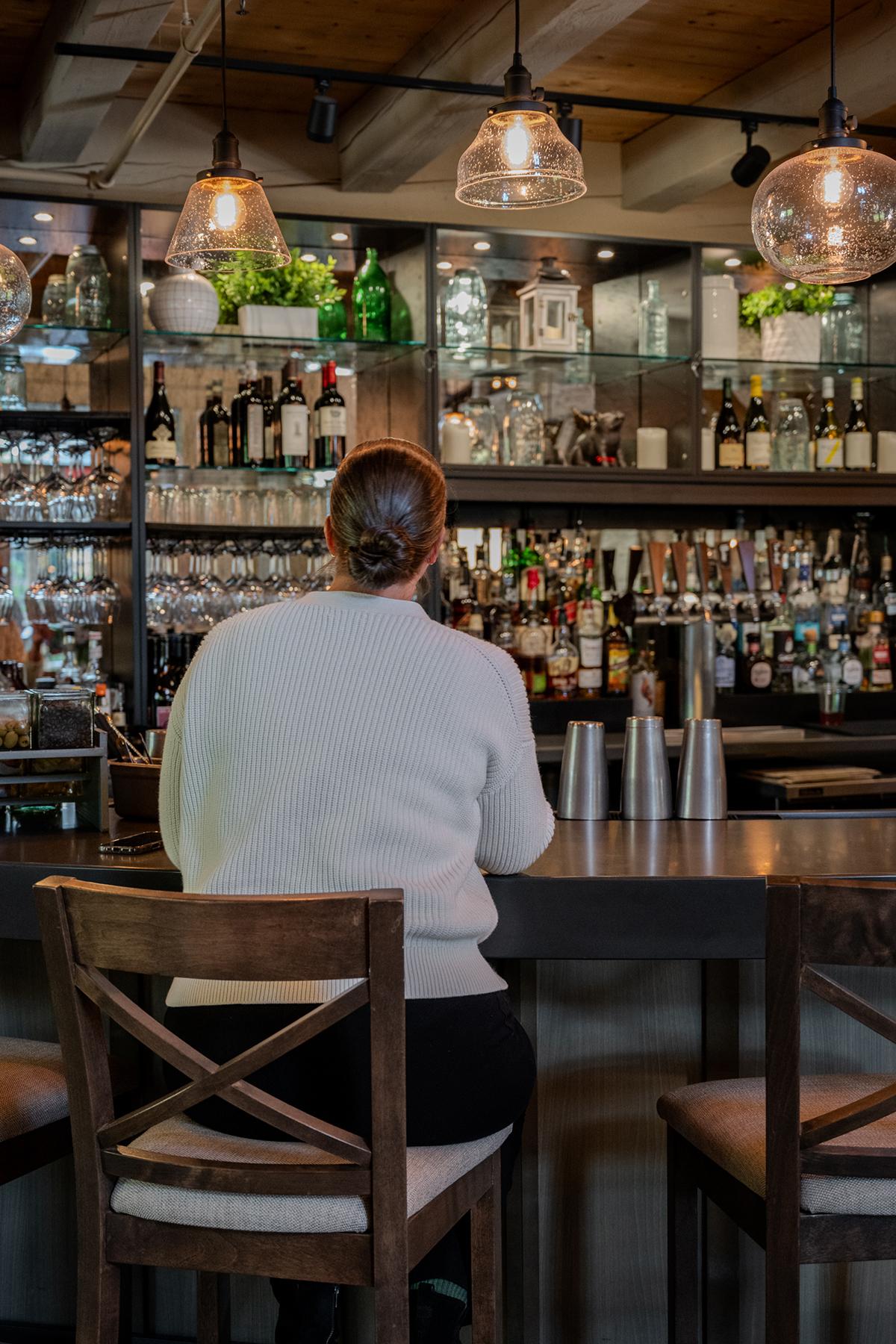 Woman seated at a bar facing shelves of bottles and hanging lights.
