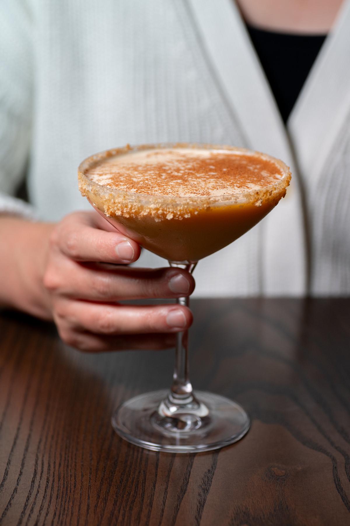 Person holding a frothy orange cocktail in a coupe glass over a wooden table
