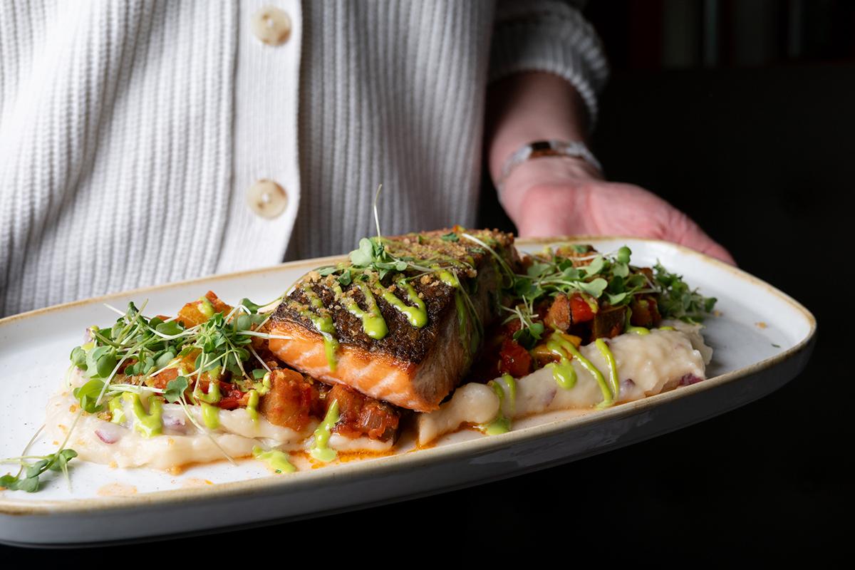 Grilled fish fillet with herbs and vegetables on a white plate, served by a chef.