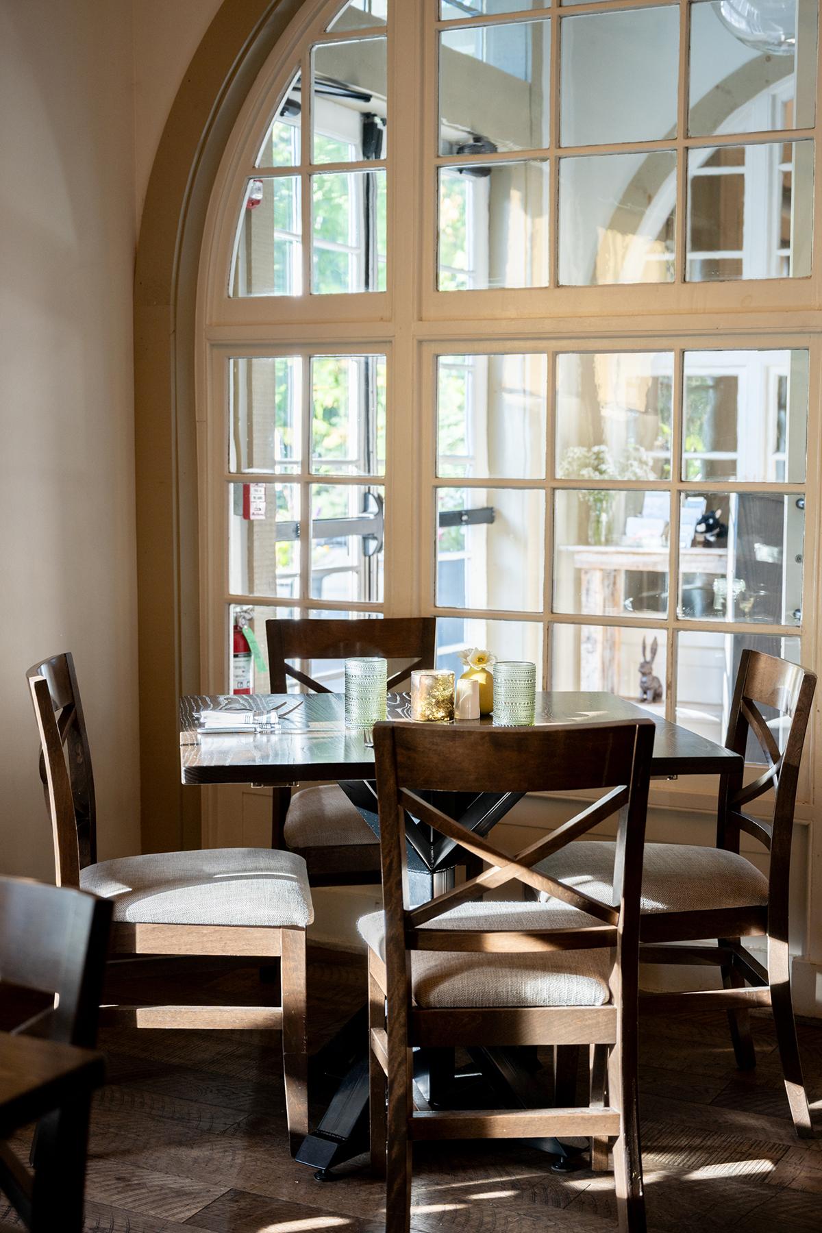 Sunlit dining area with wooden chairs and a round table by tall arched windows.